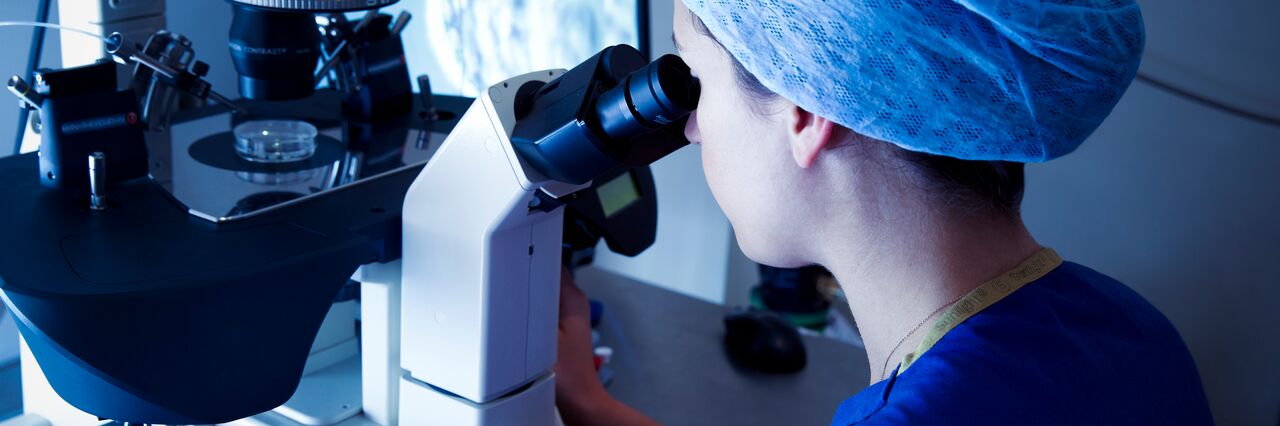 Embryologist in foreground looking down microscope at embryo