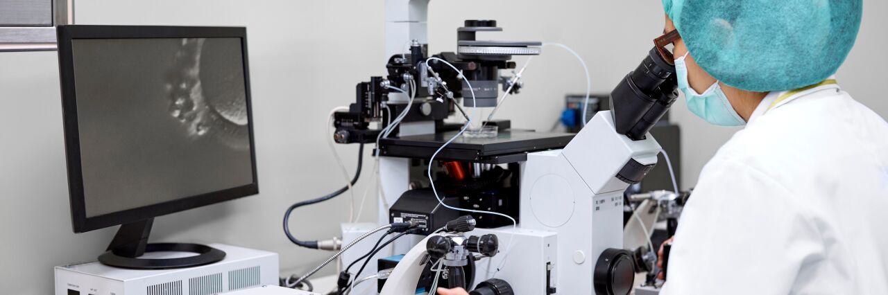 embryologist in white jacket and blue hair net looking down a microscope in lab 