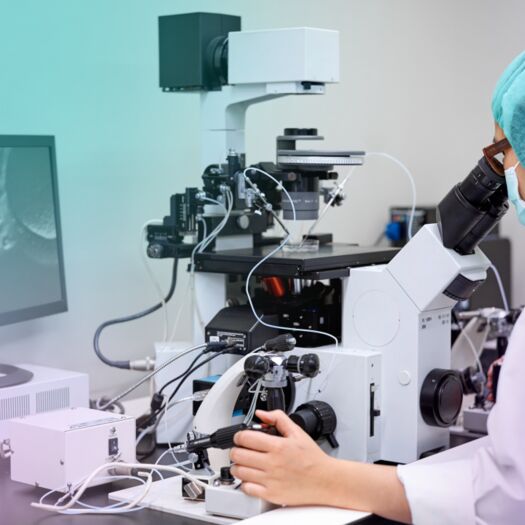 embryologist in white jacket and blue hair net looking down a microscope in lab 