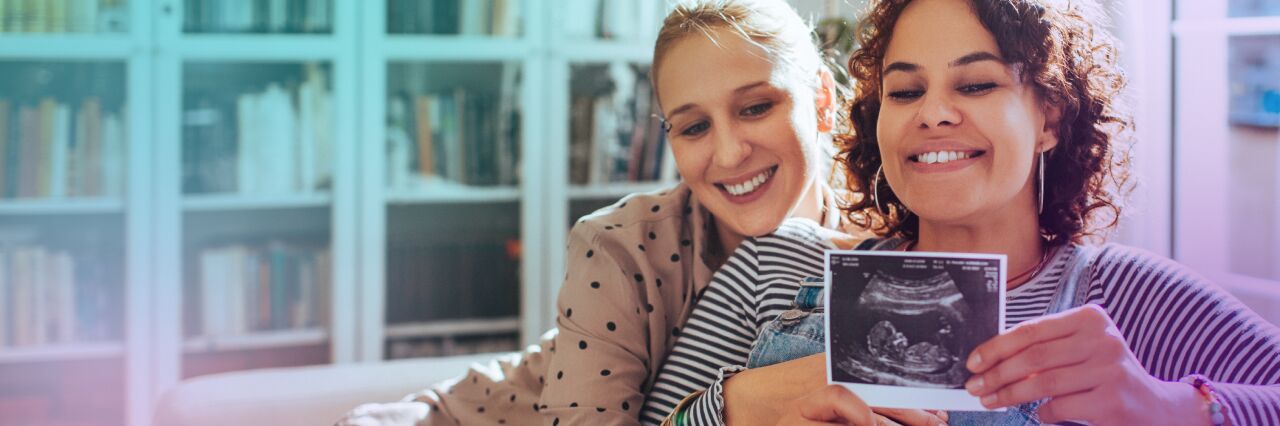 White same sex female couple hugging whilst they hold an ultrasound scan up